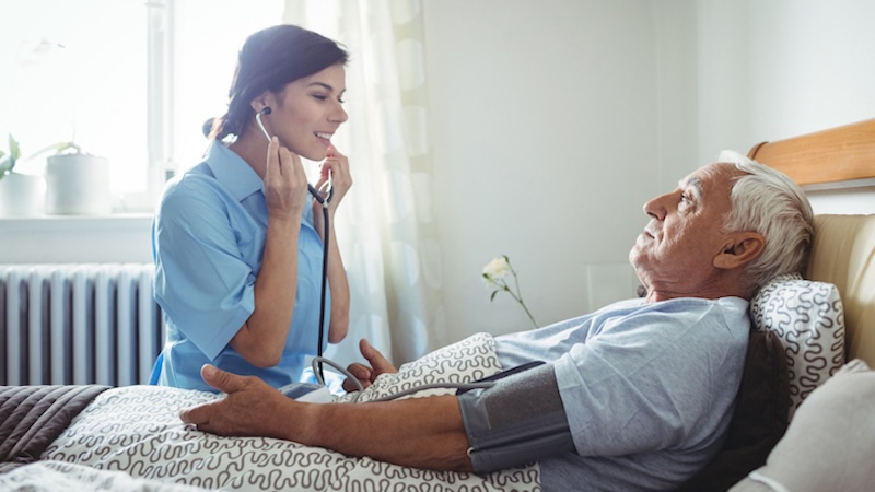 nurse sat taking the heartbeat and blood pressure of resident laying in bed